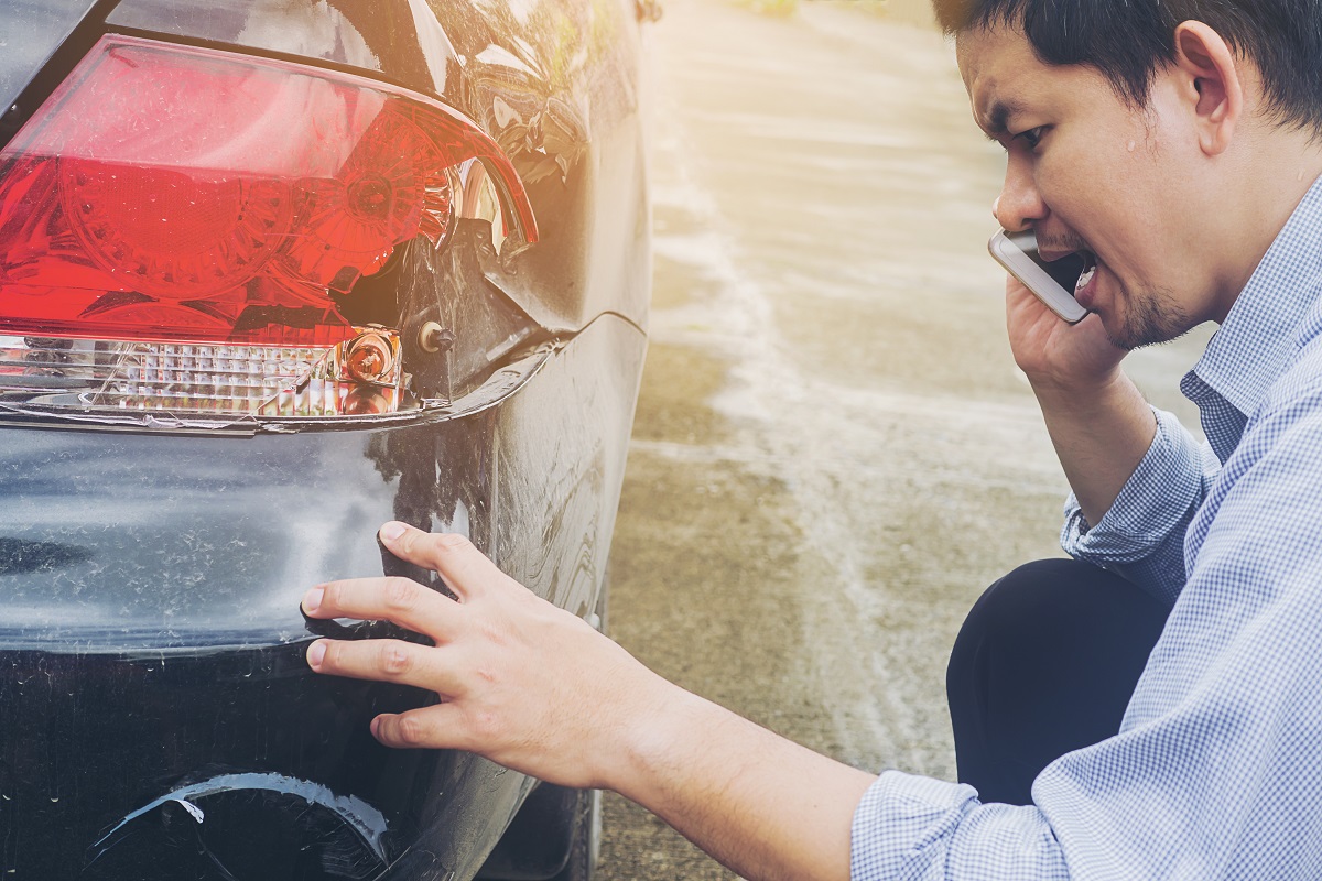man talking on a smartphone while examining a dent on the back of his car